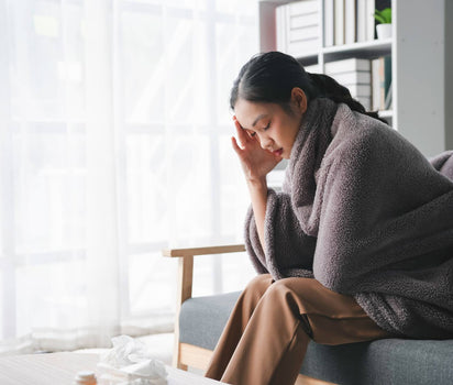 Woman wrapped in blanket on sofa with head in hand — daily multivitamin supports energy and micronutrient gaps