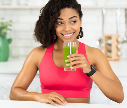 Smiling woman in pink sports top drinking green smoothie in kitchen — daily multivitamin and omega-3 support immune resilience