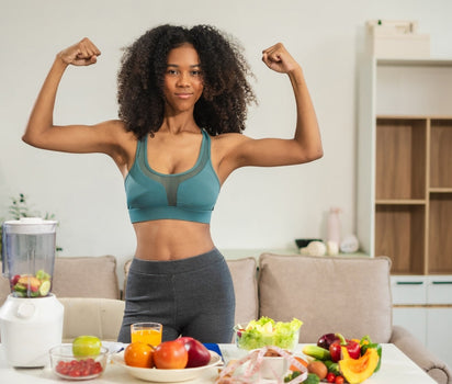 Woman flexing arms behind table of fresh fruit, vegetables and blender — probiotics support gut health and digestion