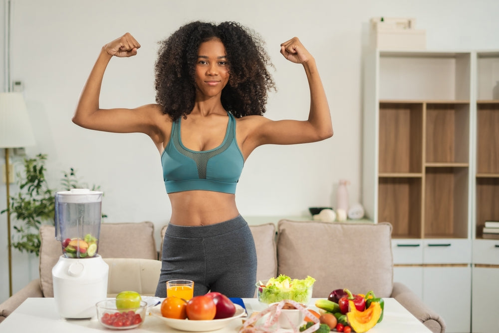Woman flexing arms behind table of fresh fruit, vegetables and blender — probiotics support gut health and digestion