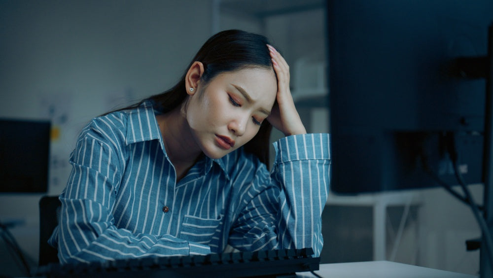 Magnesium and multivitamin support mood balance and stress — woman in striped shirt with head in hand at desk